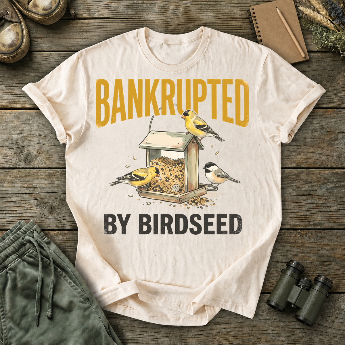 Bankrupted by Birdseed Backyard Birds Tee in Ivory, centered front design on a birdwatching flat-lay mockup