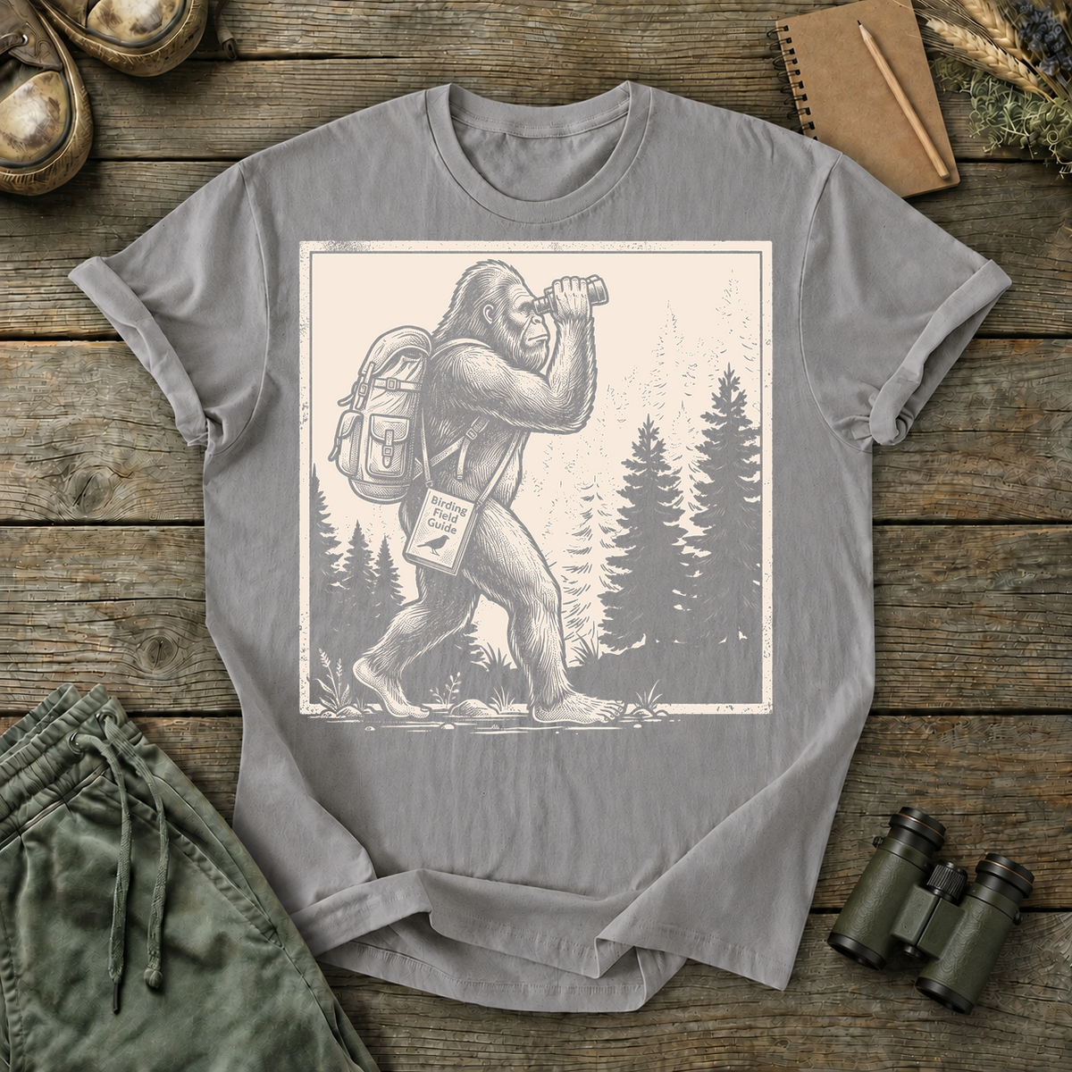Bigfoot Birdwatching Field Guide Tee in Grey, centered front design on a birdwatching flat-lay mockup