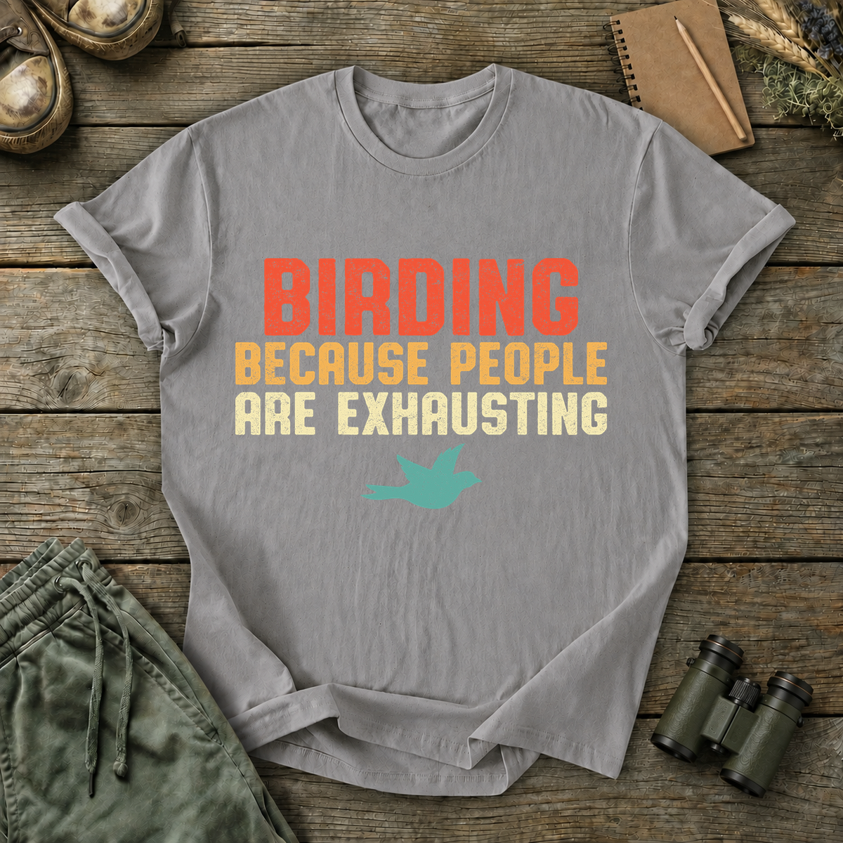 Birding Because People Are Exhausting Tee in Grey, centered front design on a birdwatching flat-lay mockup