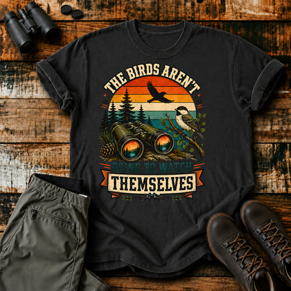 Birds Aren't Going to Watch Themselves Field Tee in Black, centered front design on a birdwatching flat-lay mockup