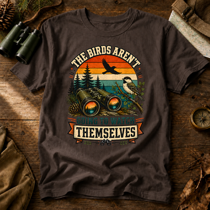 Birds Aren't Going to Watch Themselves Field Tee in Espresso, centered front design on a birdwatching flat-lay mockup