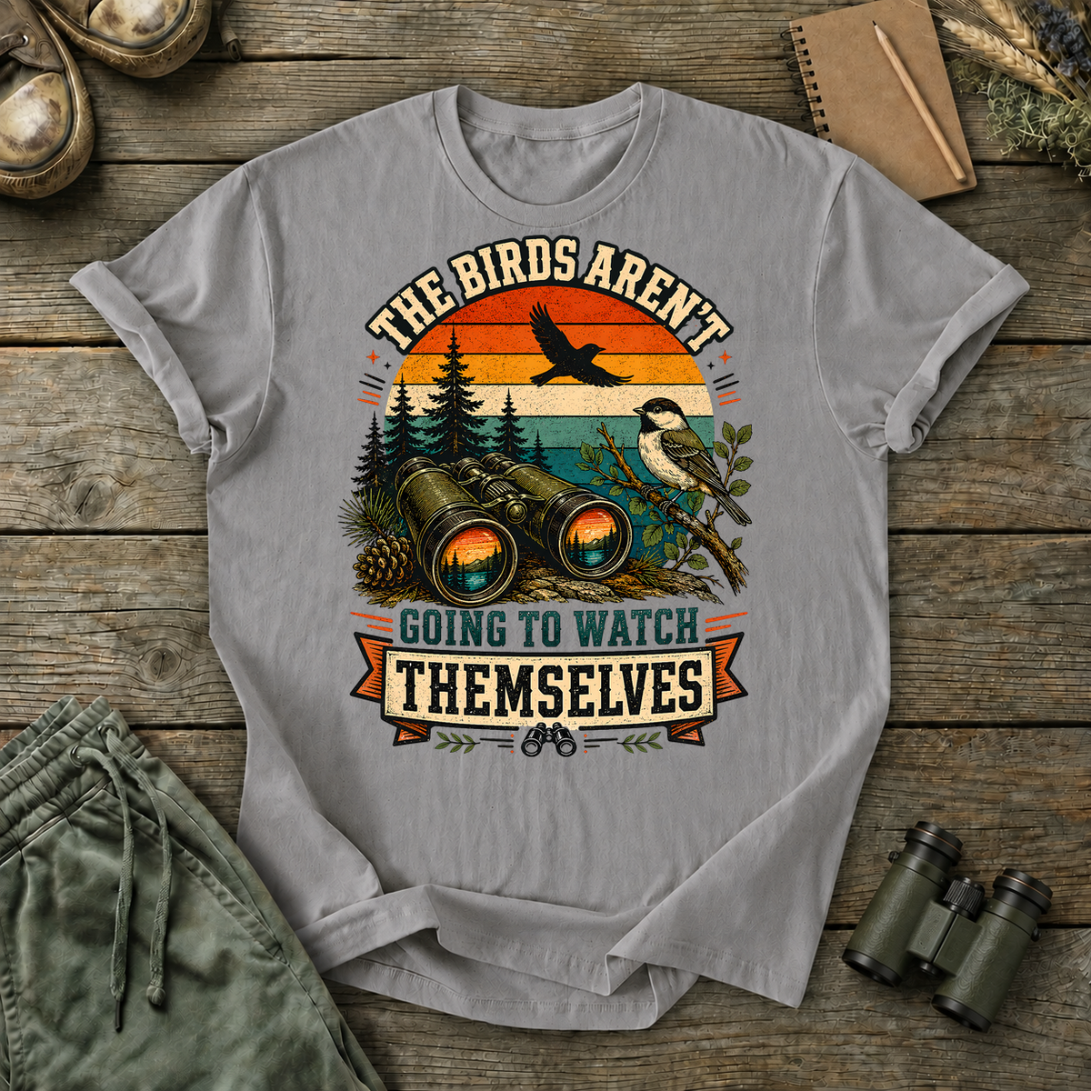 Birds Aren't Going to Watch Themselves Field Tee in Grey, centered front design on a birdwatching flat-lay mockup