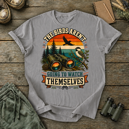 Birds Aren't Going to Watch Themselves Field Tee in Grey, centered front design on a birdwatching flat-lay mockup