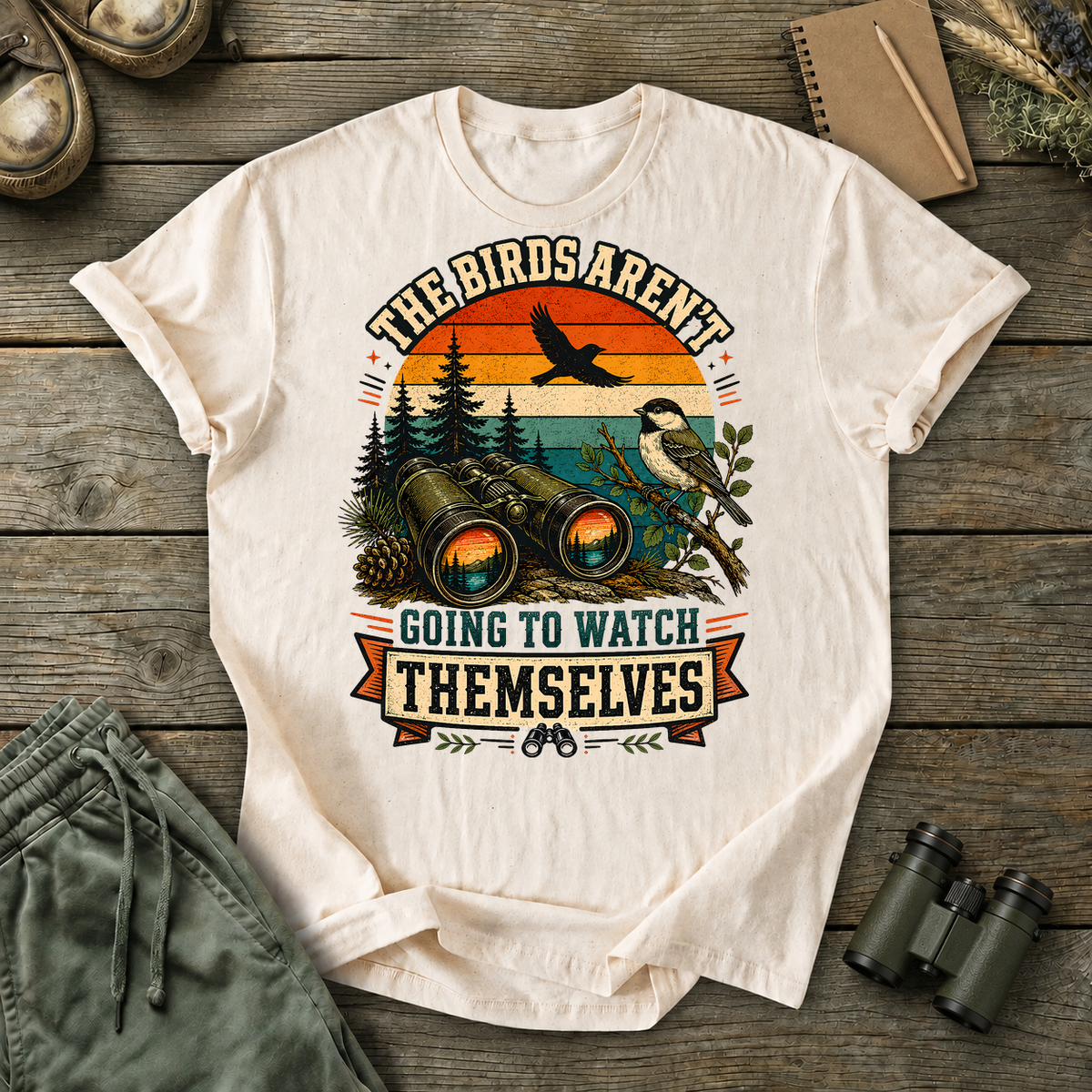 Birds Aren't Going to Watch Themselves Field Tee in Ivory, centered front design on a birdwatching flat-lay mockup