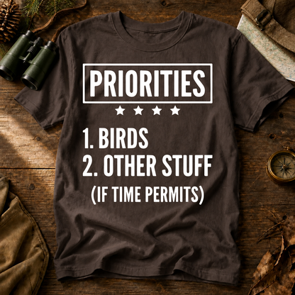 Birds First Priorities Tee in Espresso, centered front design on a birdwatching flat-lay mockup