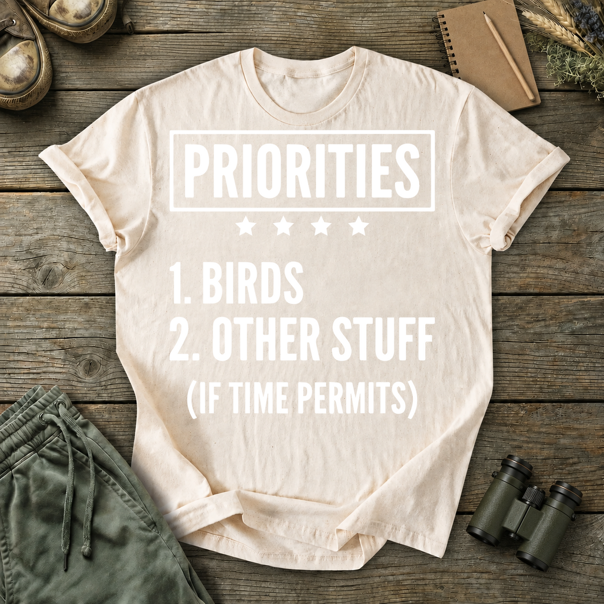 Birds First Priorities Tee in Ivory, centered front design on a birdwatching flat-lay mockup