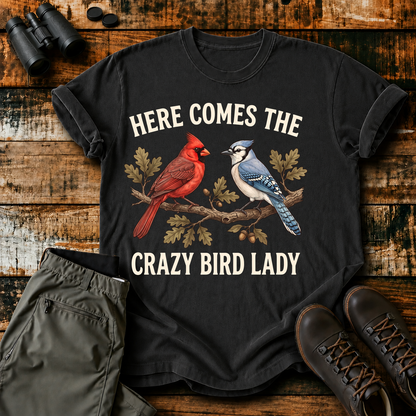 Here Comes the Crazy Bird Lady Tee in Black, centered front design on a birdwatching flat-lay mockup