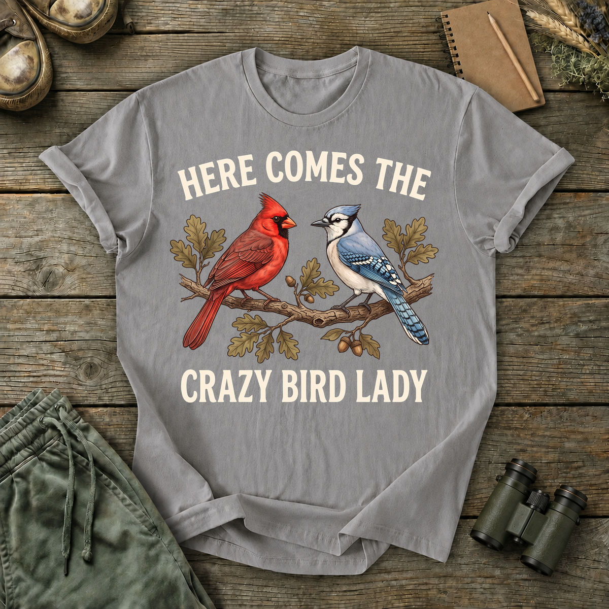 Here Comes the Crazy Bird Lady Tee in Grey, centered front design on a birdwatching flat-lay mockup