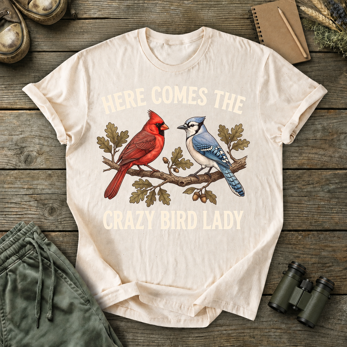 Here Comes the Crazy Bird Lady Tee in Ivory, centered front design on a birdwatching flat-lay mockup