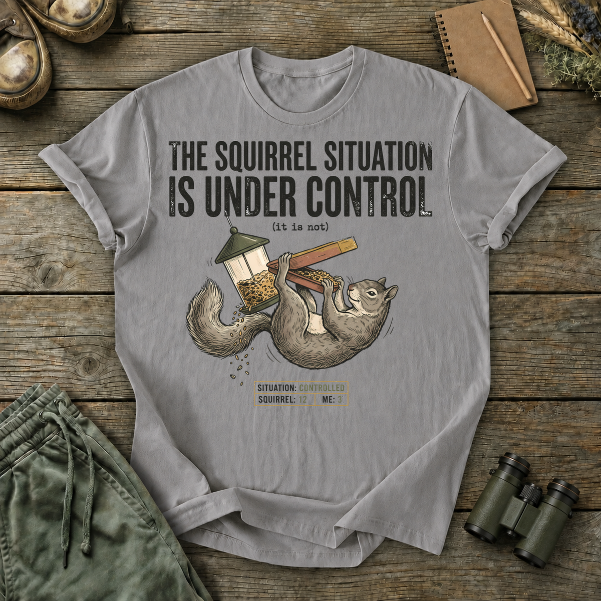 Squirrel Situation Under Control Tee in Grey, centered front design on a birdwatching flat-lay mockup