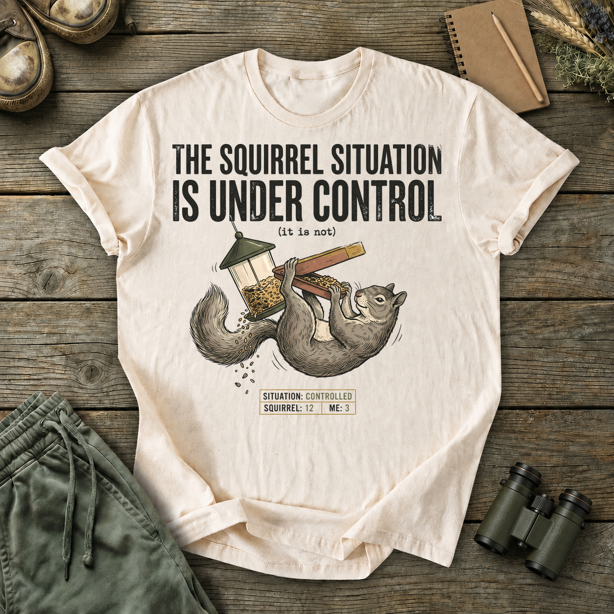 Squirrel Situation Under Control Tee in Ivory, centered front design on a birdwatching flat-lay mockup