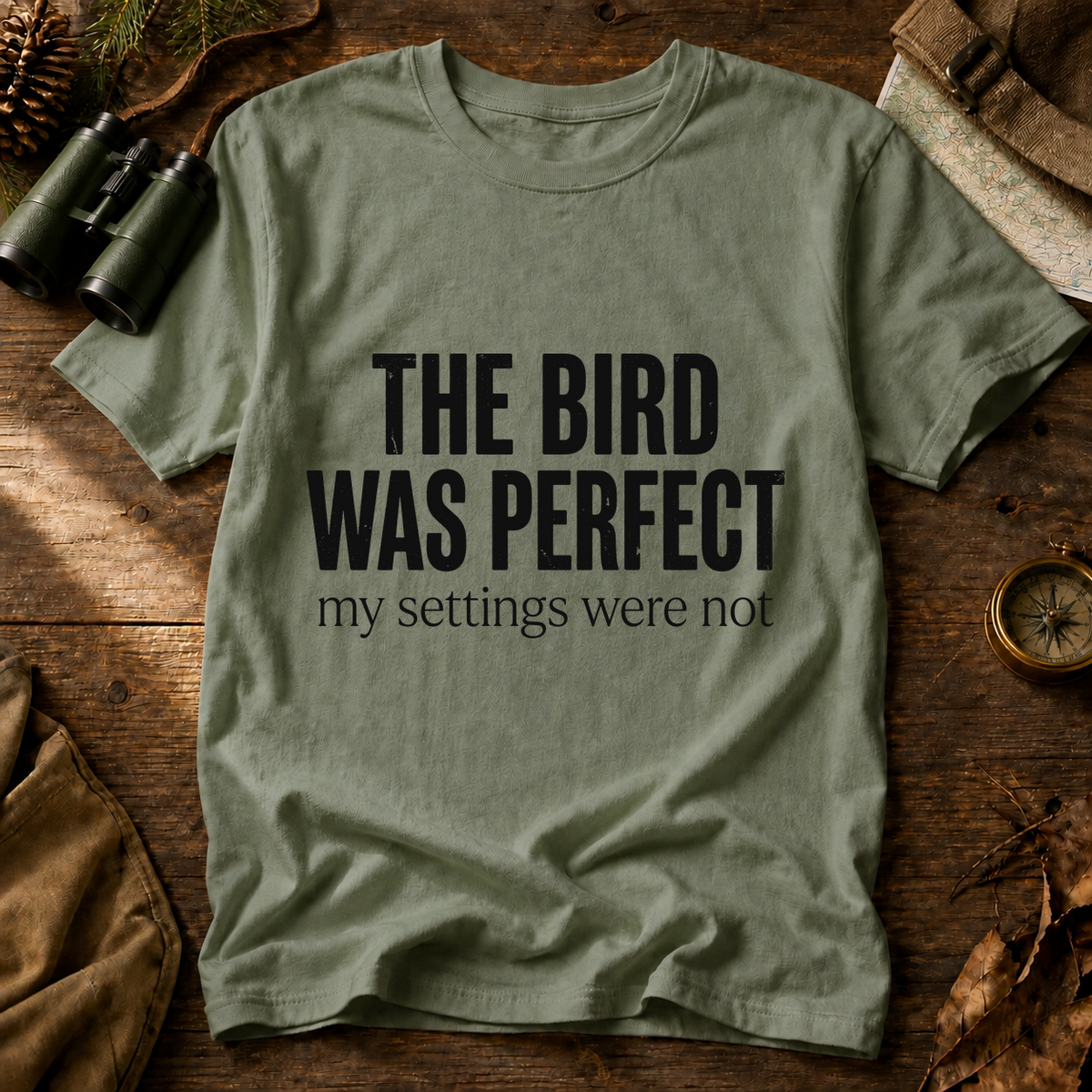 The Bird Was Perfect My Settings Were Not Tee in Bay, centered front design on a birdwatching flat-lay mockup