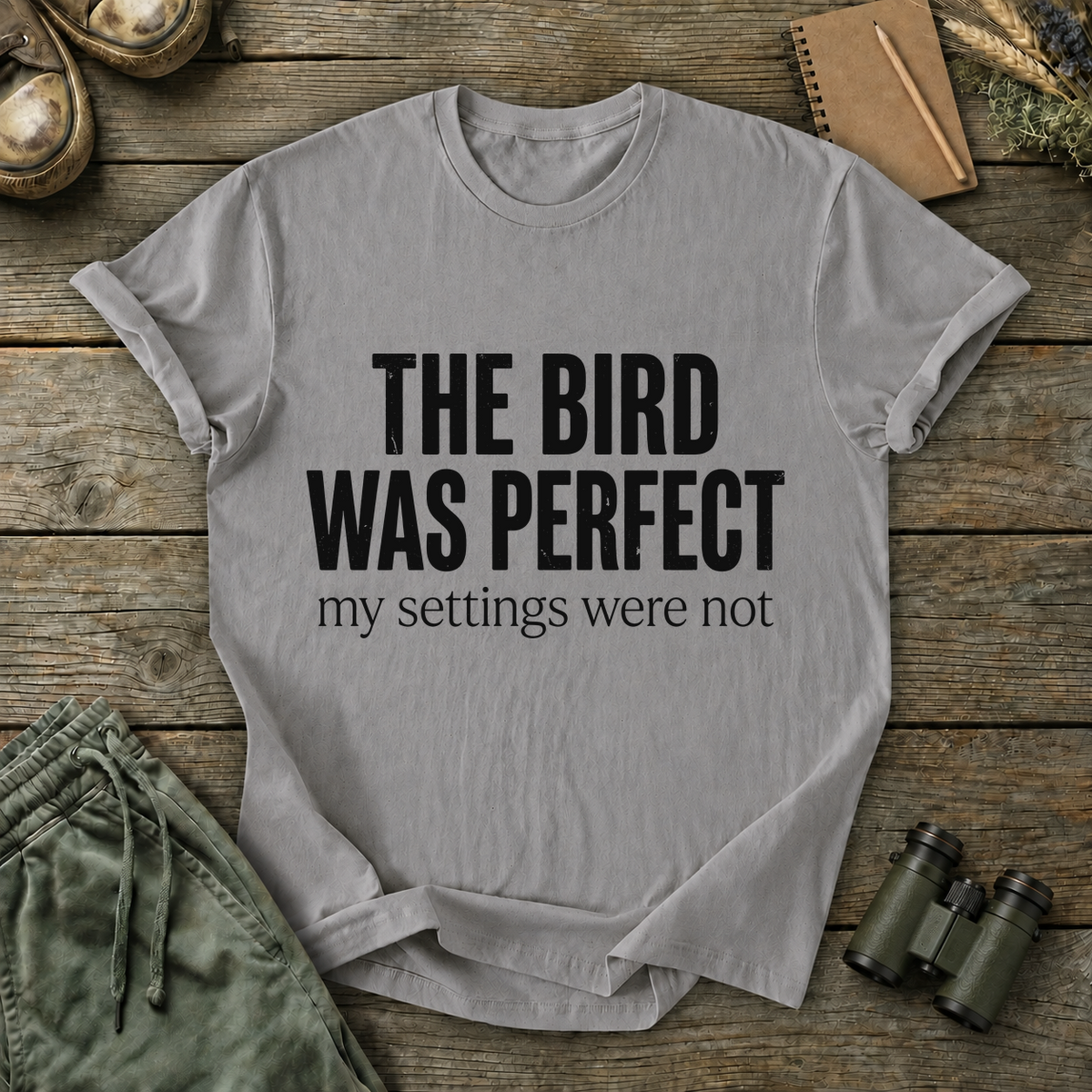 The Bird Was Perfect My Settings Were Not Tee in Grey, centered front design on a birdwatching flat-lay mockup