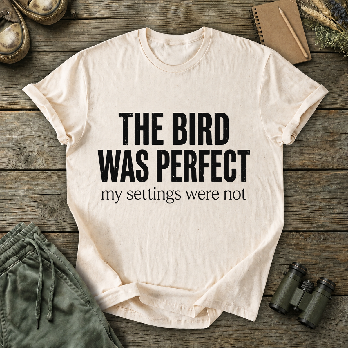 The Bird Was Perfect My Settings Were Not Tee in Ivory, centered front design on a birdwatching flat-lay mockup