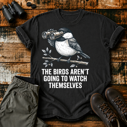 The Birds Aren't Going to Watch Themselves Tee in Black, centered front design on a birdwatching flat-lay mockup
