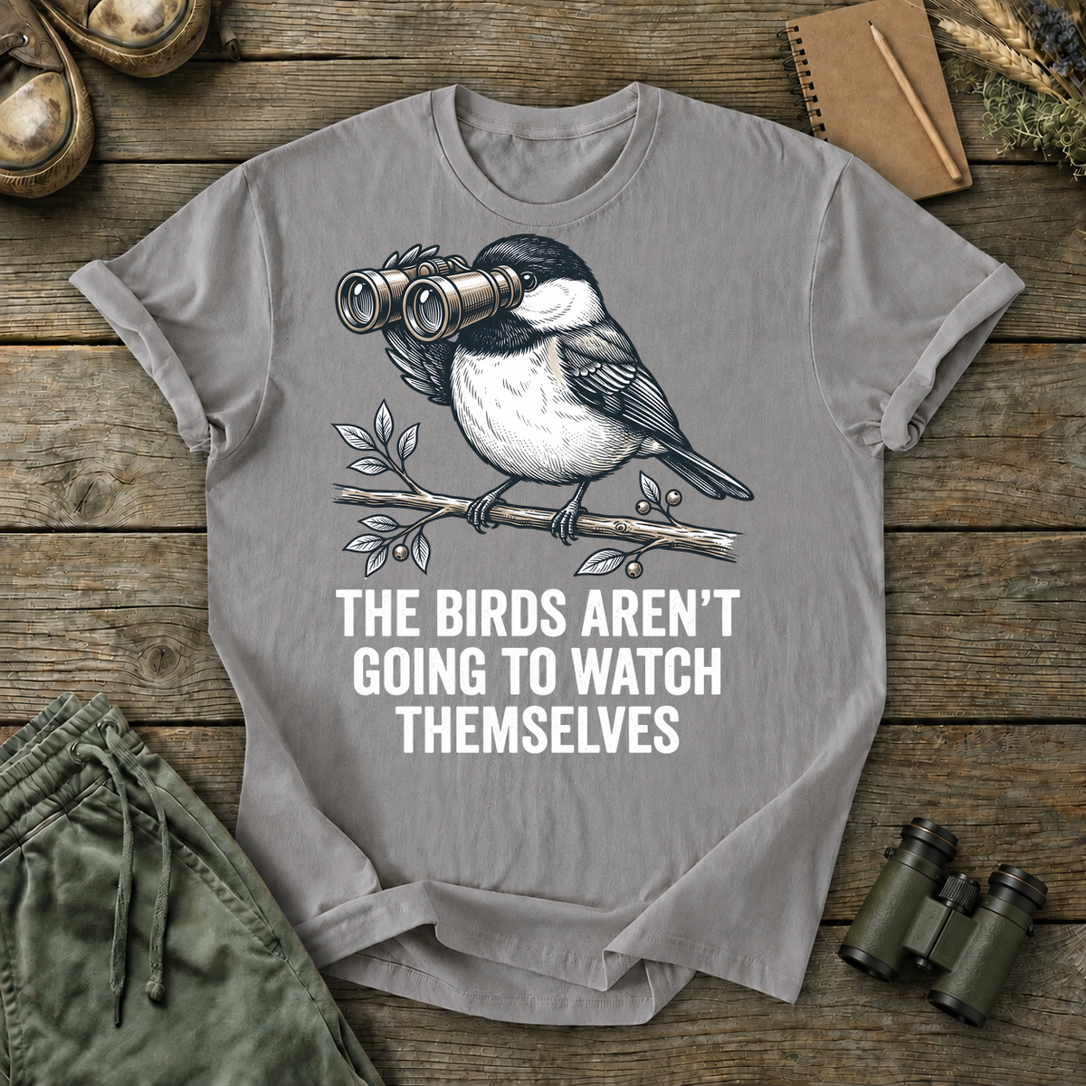 The Birds Aren't Going to Watch Themselves Tee in Grey, centered front design on a birdwatching flat-lay mockup