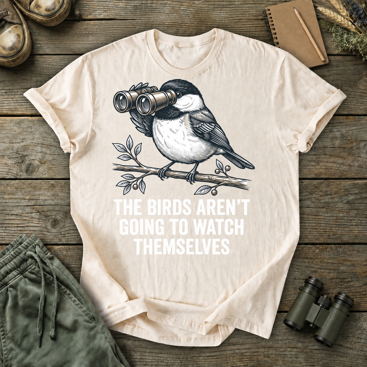 The Birds Aren't Going to Watch Themselves Tee in Ivory, centered front design on a birdwatching flat-lay mockup