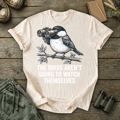 The Birds Aren't Going to Watch Themselves Tee in Ivory, centered front design on a birdwatching flat-lay mockup