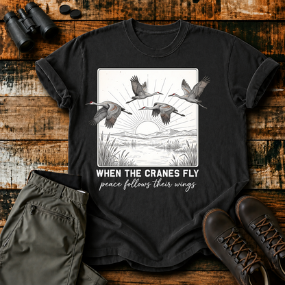 When the Cranes Fly Peace Follows Tee in Black, centered front design on a birdwatching flat-lay mockup