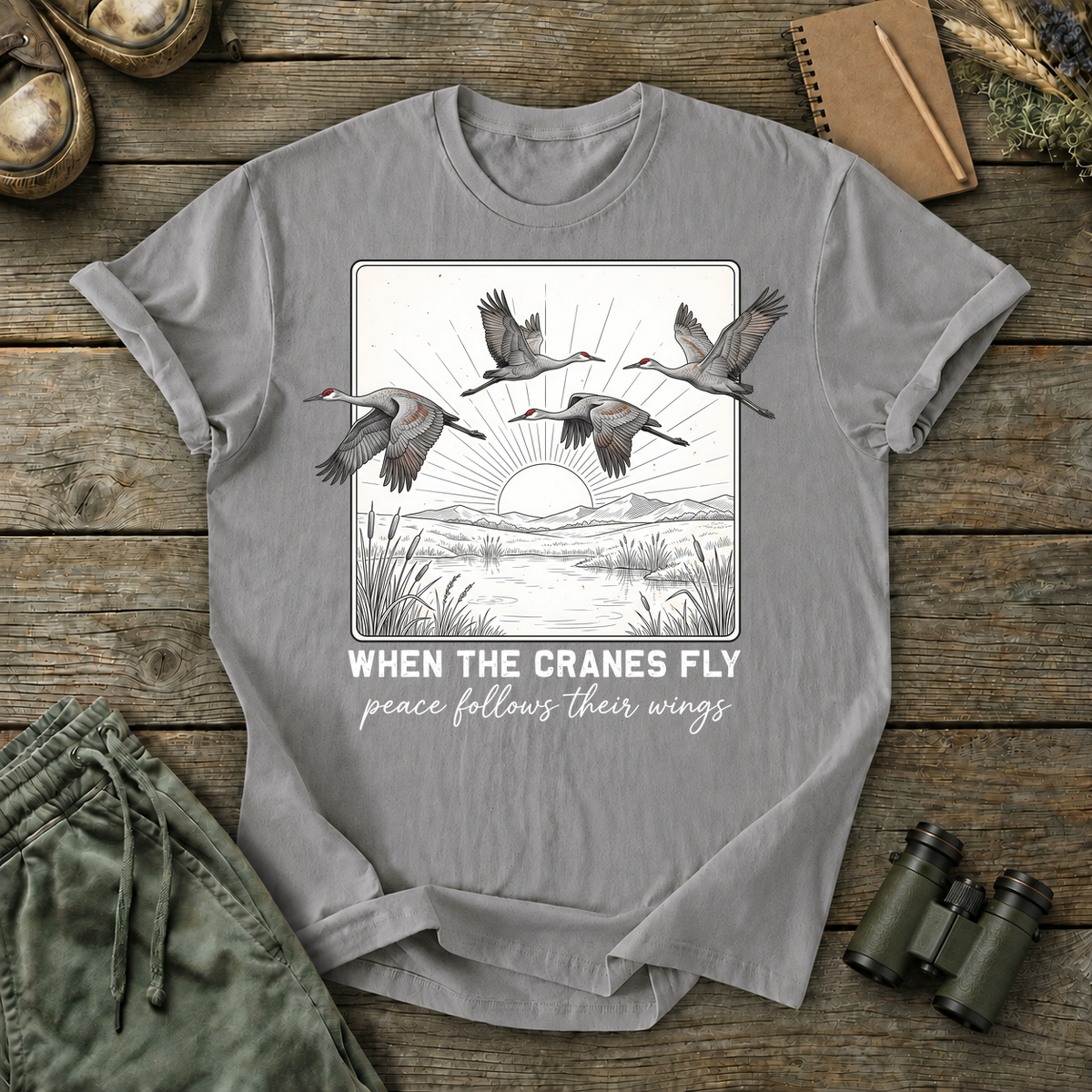 When the Cranes Fly Peace Follows Tee in Grey, centered front design on a birdwatching flat-lay mockup